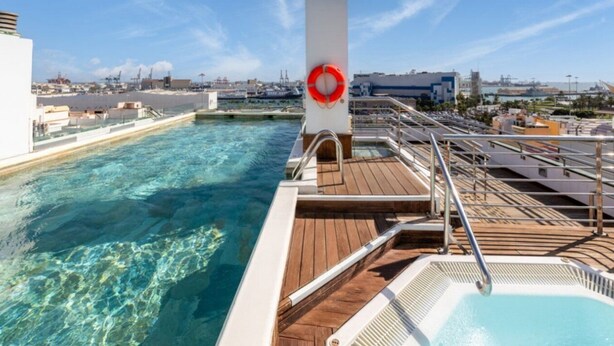 Poolbereich auf dem Deck eines Kreuzfahrtschiffs mit einem Rettungsring, klarem Wasser und Blick auf die Hafenstadt im Hintergrund.