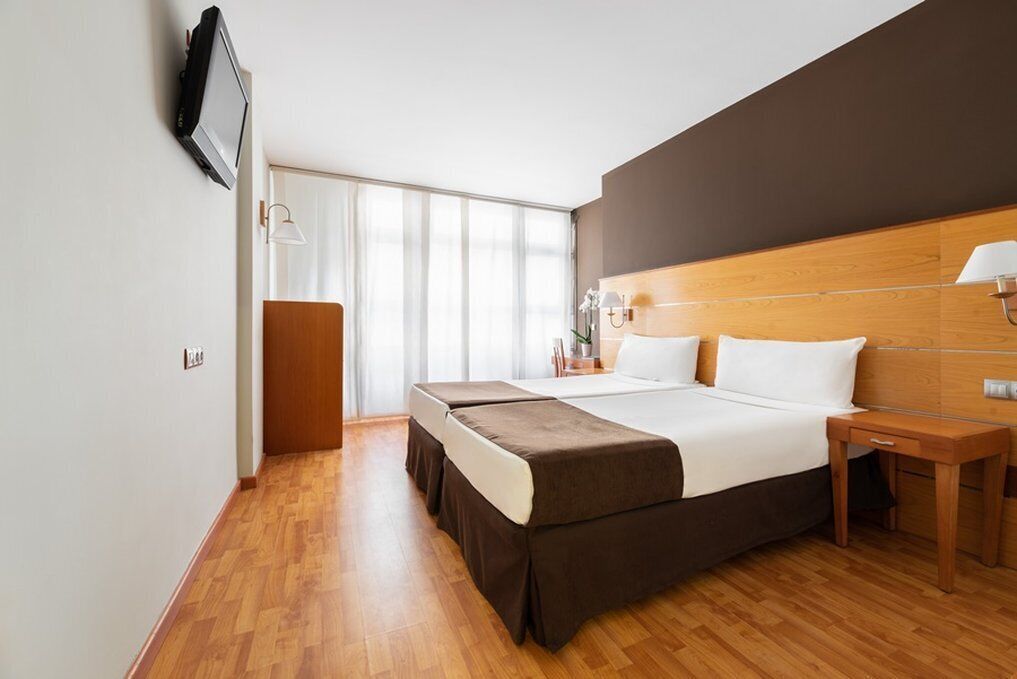 A hotel room with a large bed, wooden headboard, bedside tables, a flat-screen TV mounted on the wall, and a sliding door leading to a balcony.