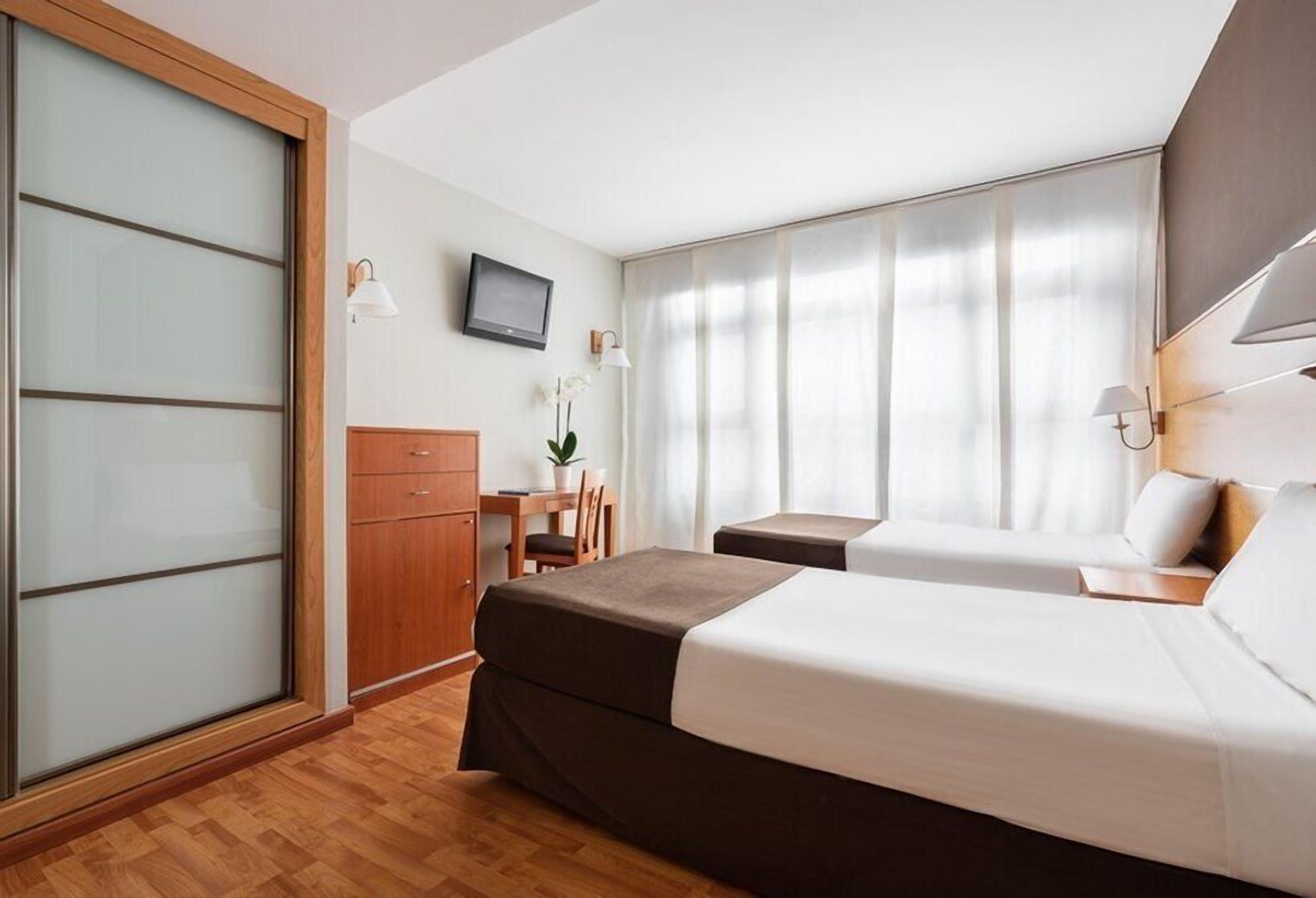 A hotel room with two beds, a desk, a chair, a TV, and a sliding door.