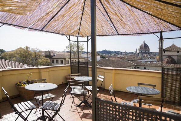 A terrace with two round tables and chairs, a potted plant, and a view of a dome and buildings in the distance.