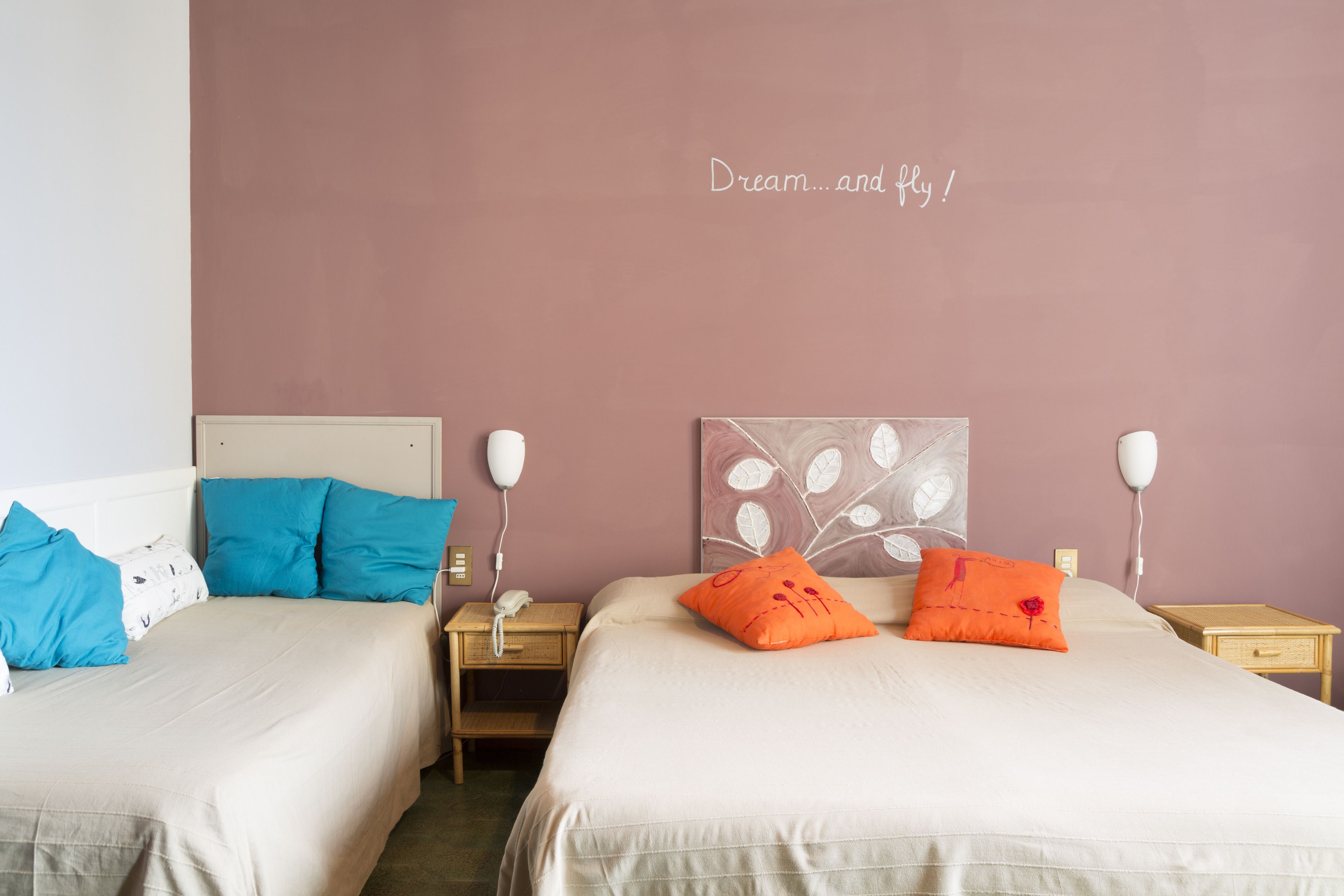 A hotel room with two beds, each with a different color pillow, a bedside table with a lamp, and a wall with a decorative leaf pattern.