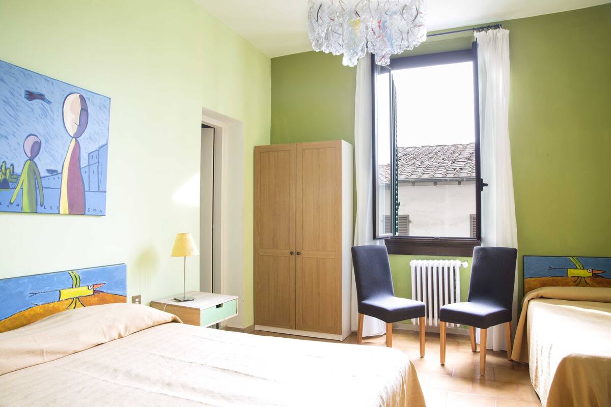 A hotel room with a bed, two chairs, a wardrobe, a radiator, and a window with curtains.