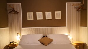 A neatly made bed with white linens and a single brown pillow, flanked by two bedside tables with lamps, in a room with a brown wall and framed pictures above the bed. - Hotel Panorama (Florence)