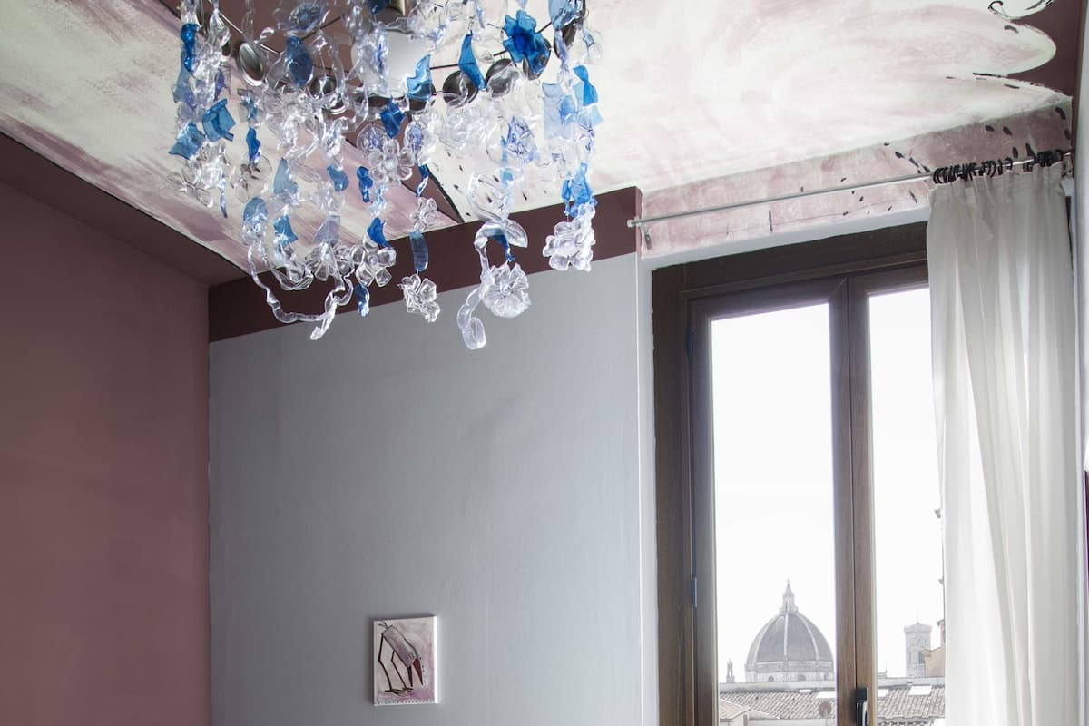 A hotel room with a bed, two chairs, a desk with a computer, a radiator, and a window with a city view.
