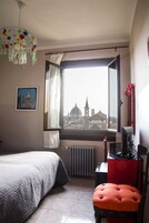 A bedroom with a bed, a chair, a desk, a radiator, and a window with a view of a building.