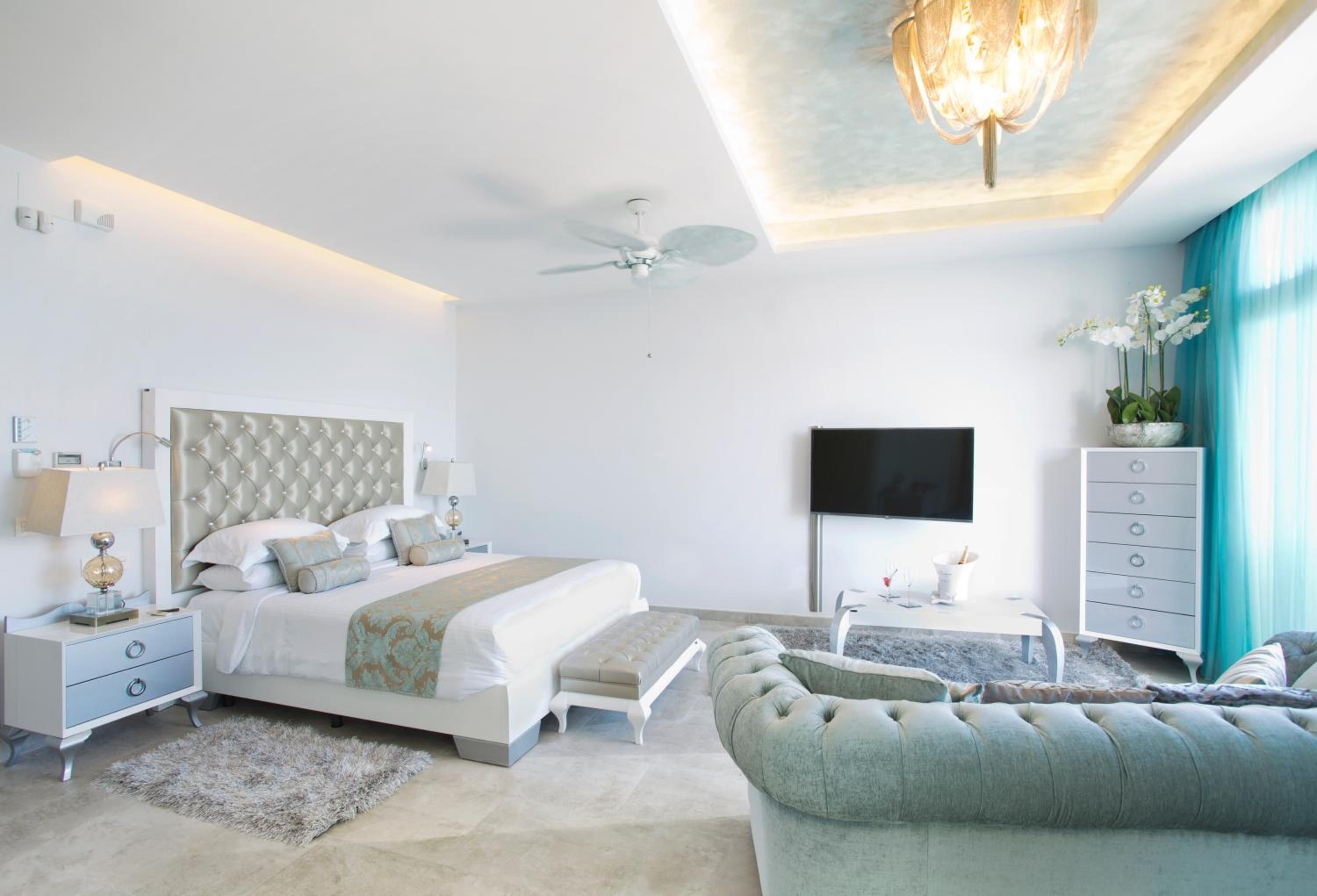 A modern bedroom with a large bed, a ceiling fan, a flat-screen TV, a dresser, and a seating area with a sofa and armchairs.