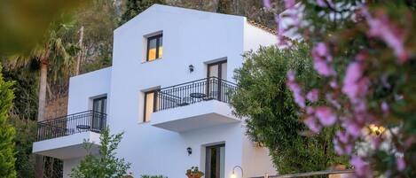 A two-story white building with balconies, surrounded by greenery and a stone wall.