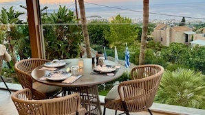 Un espacio exterior para comer con sillas de mimbre y una mesa redonda preparado para dos personas, con vistas al mar, palmeras y edificios.