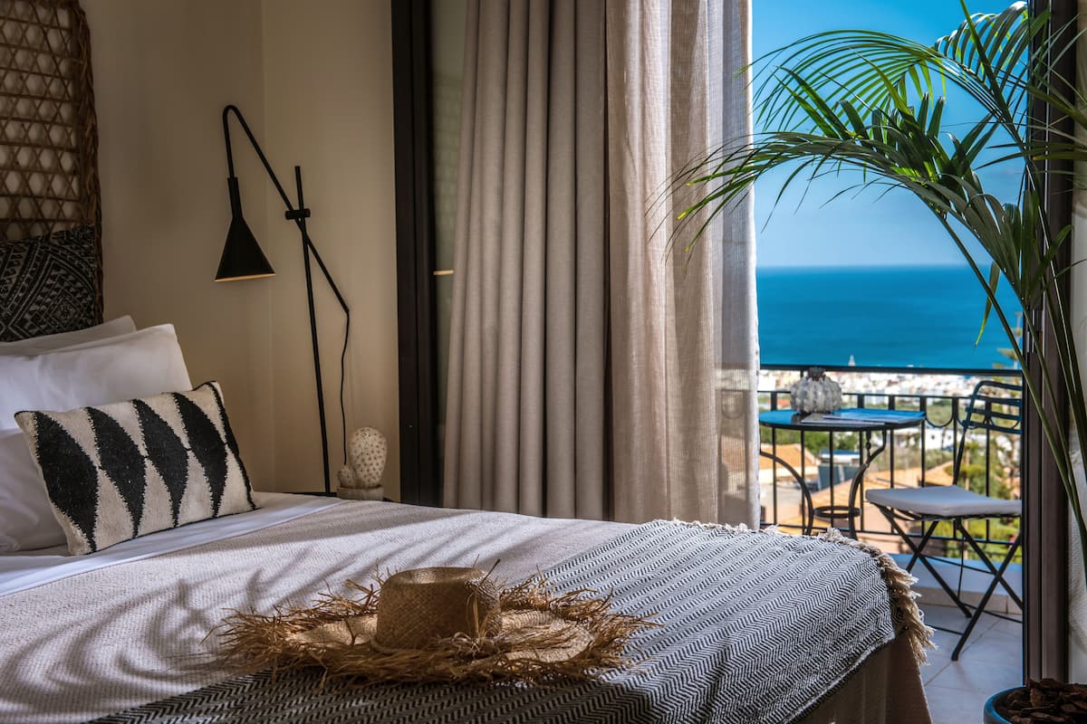 Un dormitorio con cama, lámpara de noche, vista al océano y balcón con mesa y sillas.