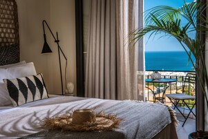 A bedroom with a bed, a bedside lamp, a view of the ocean, and a balcony with a table and chairs.