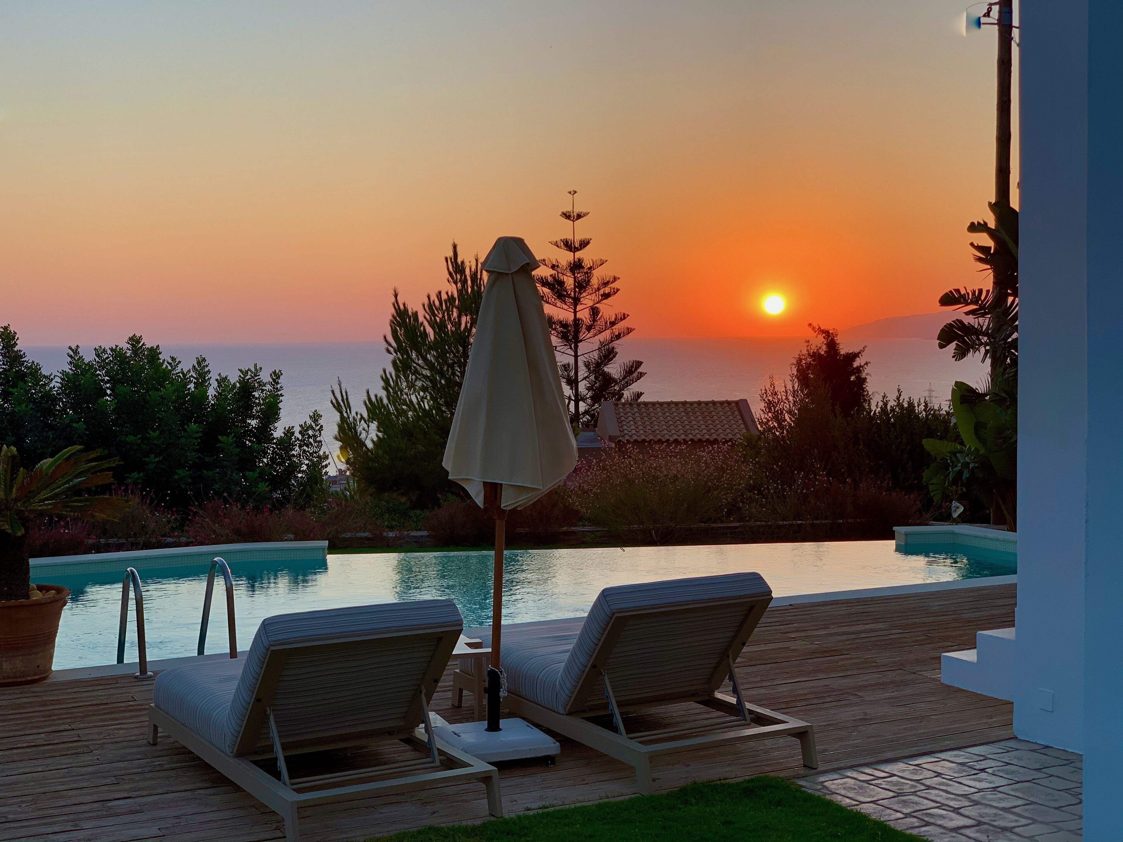 A wooden deck with two lounge chairs and a table, overlooking a swimming pool and a sunset.