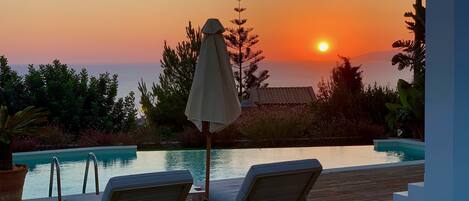 A wooden deck with two lounge chairs and a table, overlooking a swimming pool and a sunset.