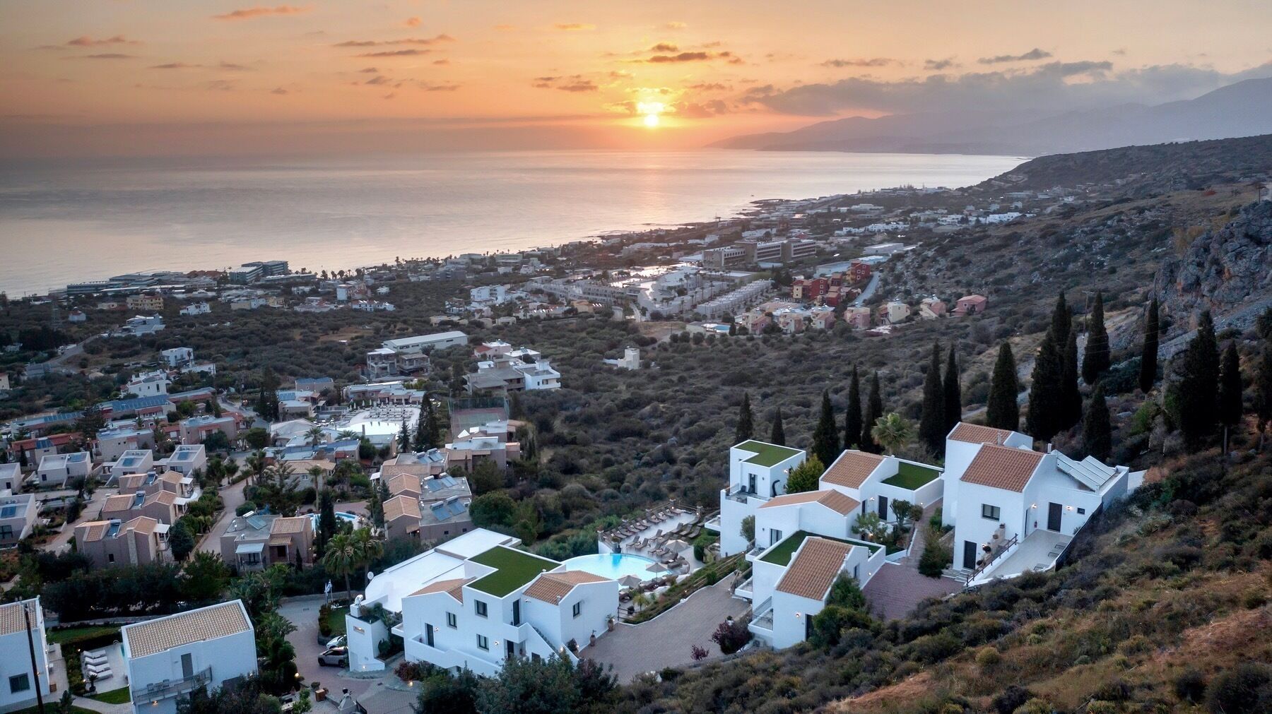 A coastal town with white buildings, a swimming pool, and a view of the sea at sunset.