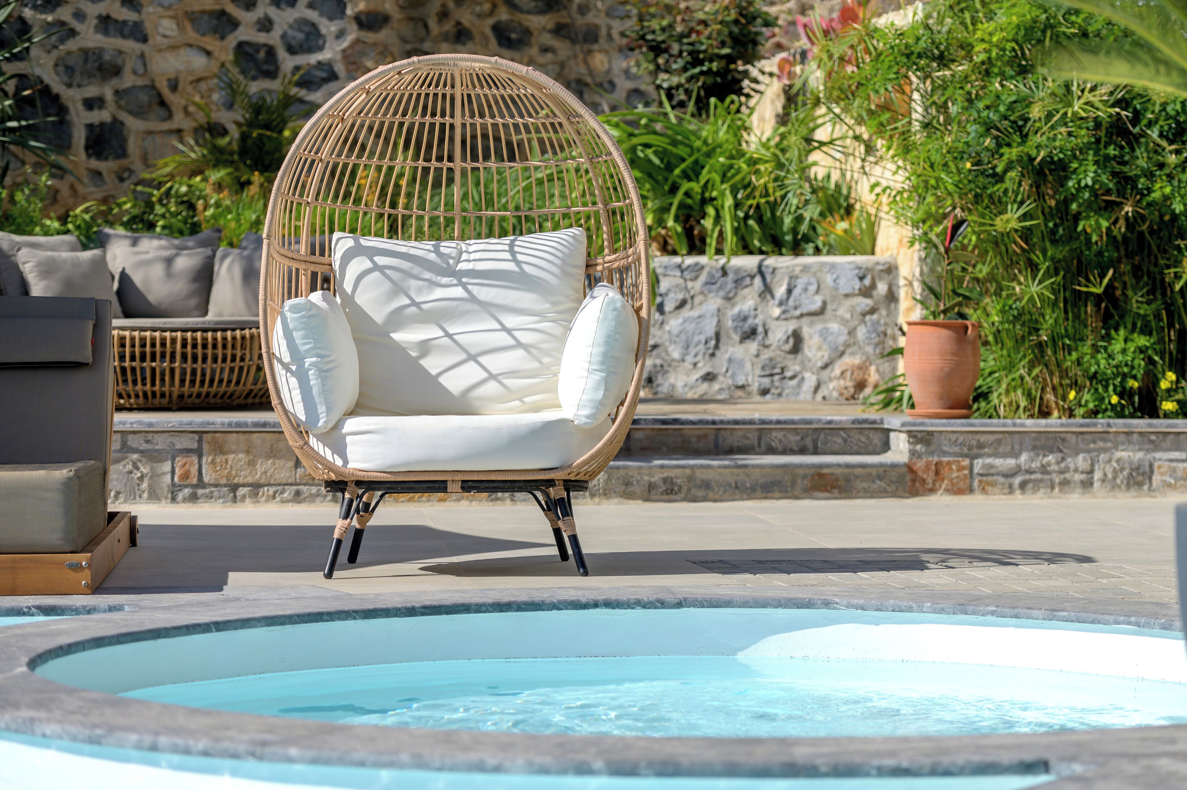 A wicker chair with white cushions by a pool, stone wall, and greenery.