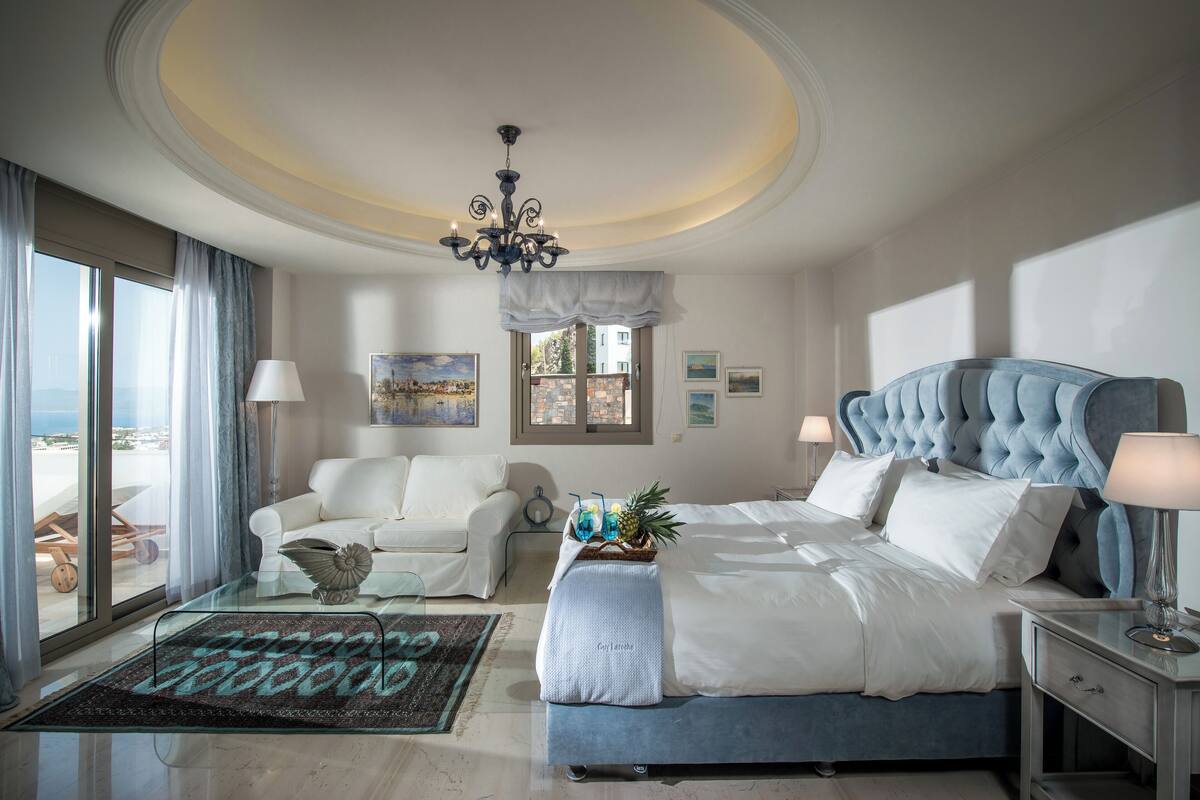 Un dormitorio amplio con una cama grande, una zona de estar con un sofá blanco y vistas a la ciudad desde las ventanas.