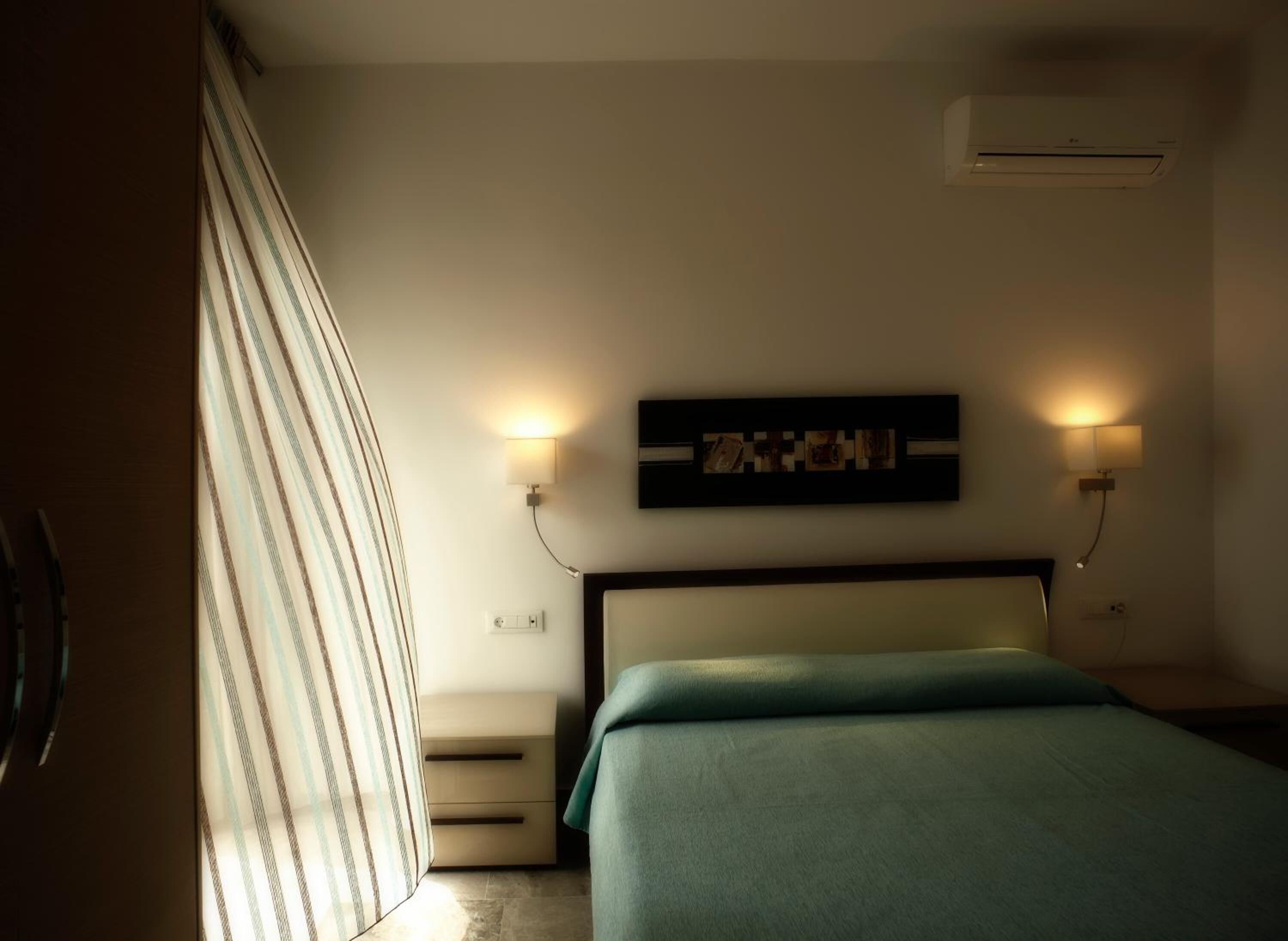 A hotel room with a bed, a nightstand, a wall-mounted air conditioner, and two wall sconces.