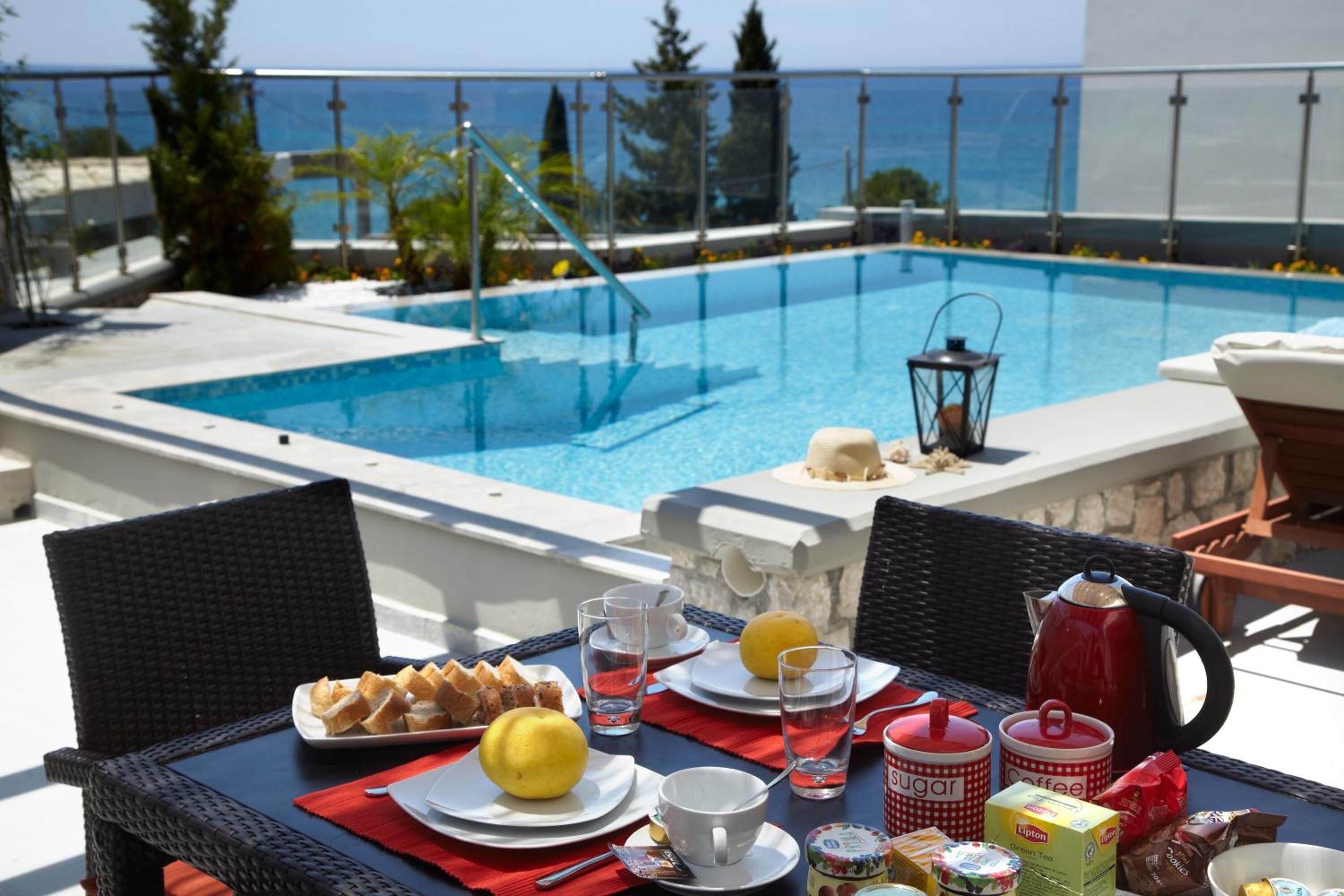 A dining table with breakfast items, a red kettle, and a view of a pool and ocean.