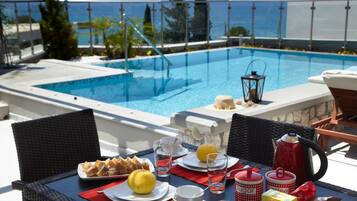 A dining table with breakfast items, a red kettle, and a view of a pool and ocean.