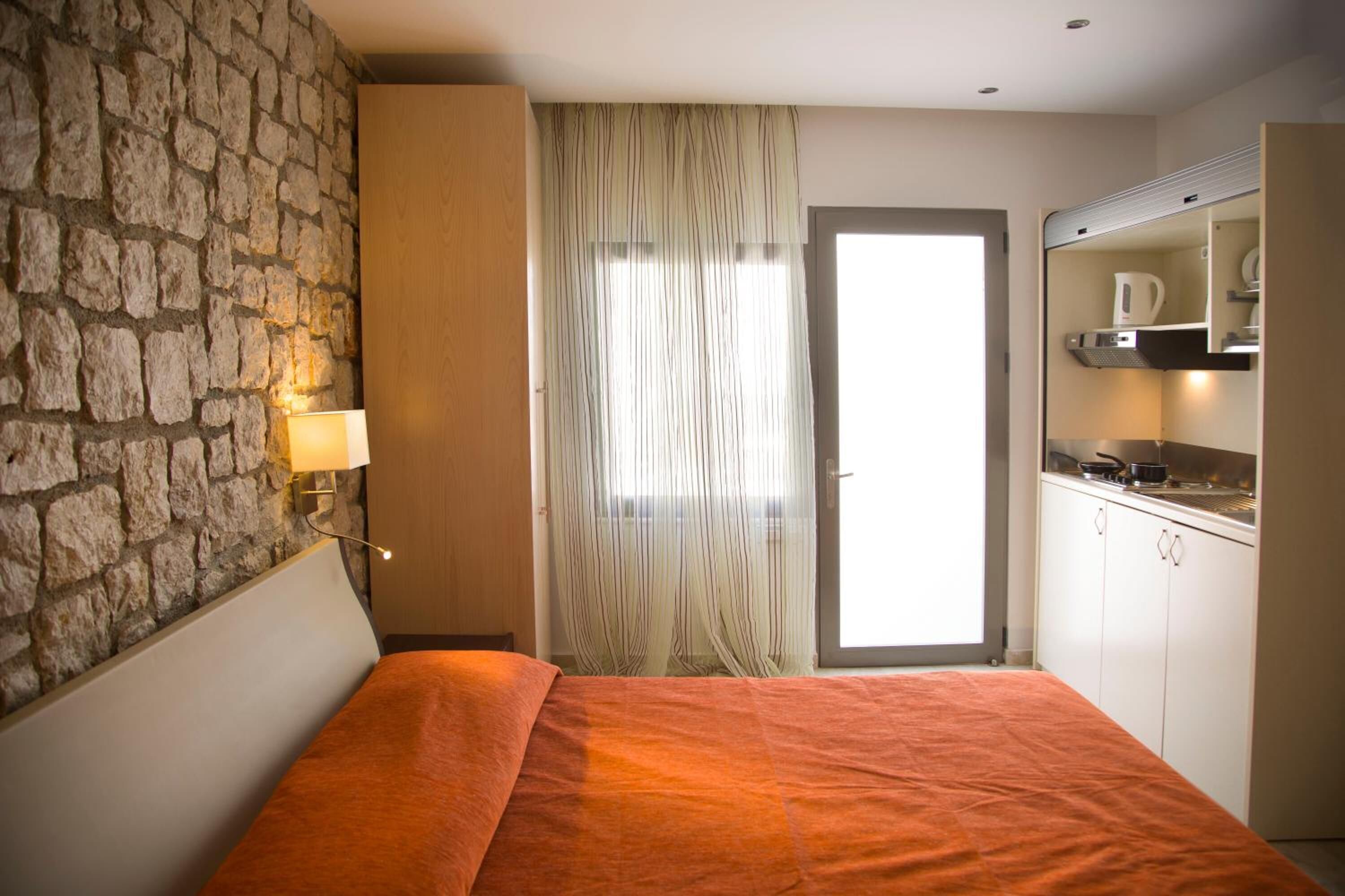 Een slaapkamer met een stenen muur, een bed met een oranje sprei, een raam met gordijnen, en een kitchenette met een waterkoker.