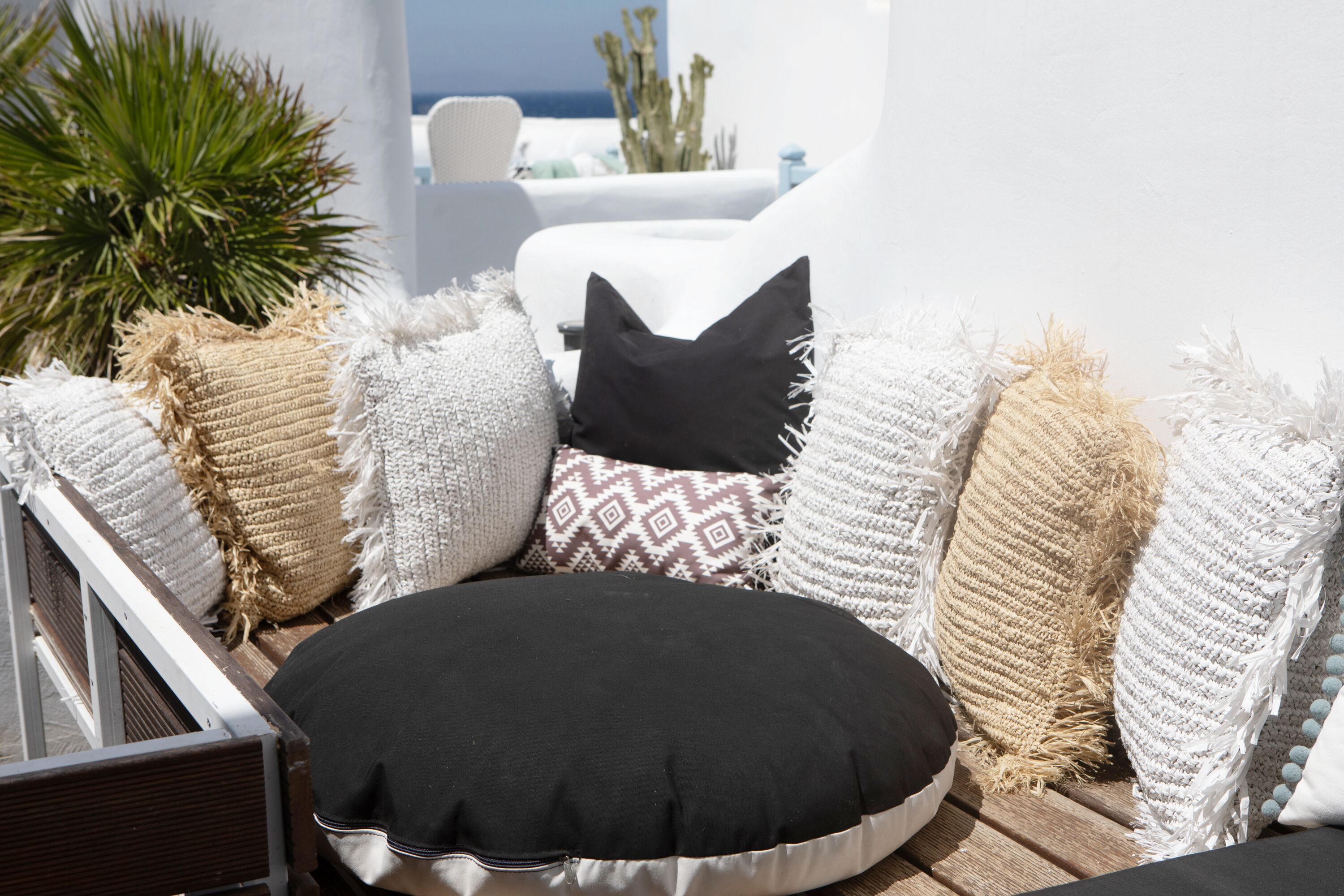 Un coin salon agrémenté de différents coussins aux textures variées et d’un coussin noir, le tout placé devant un mur blanc avec vue sur la mer.