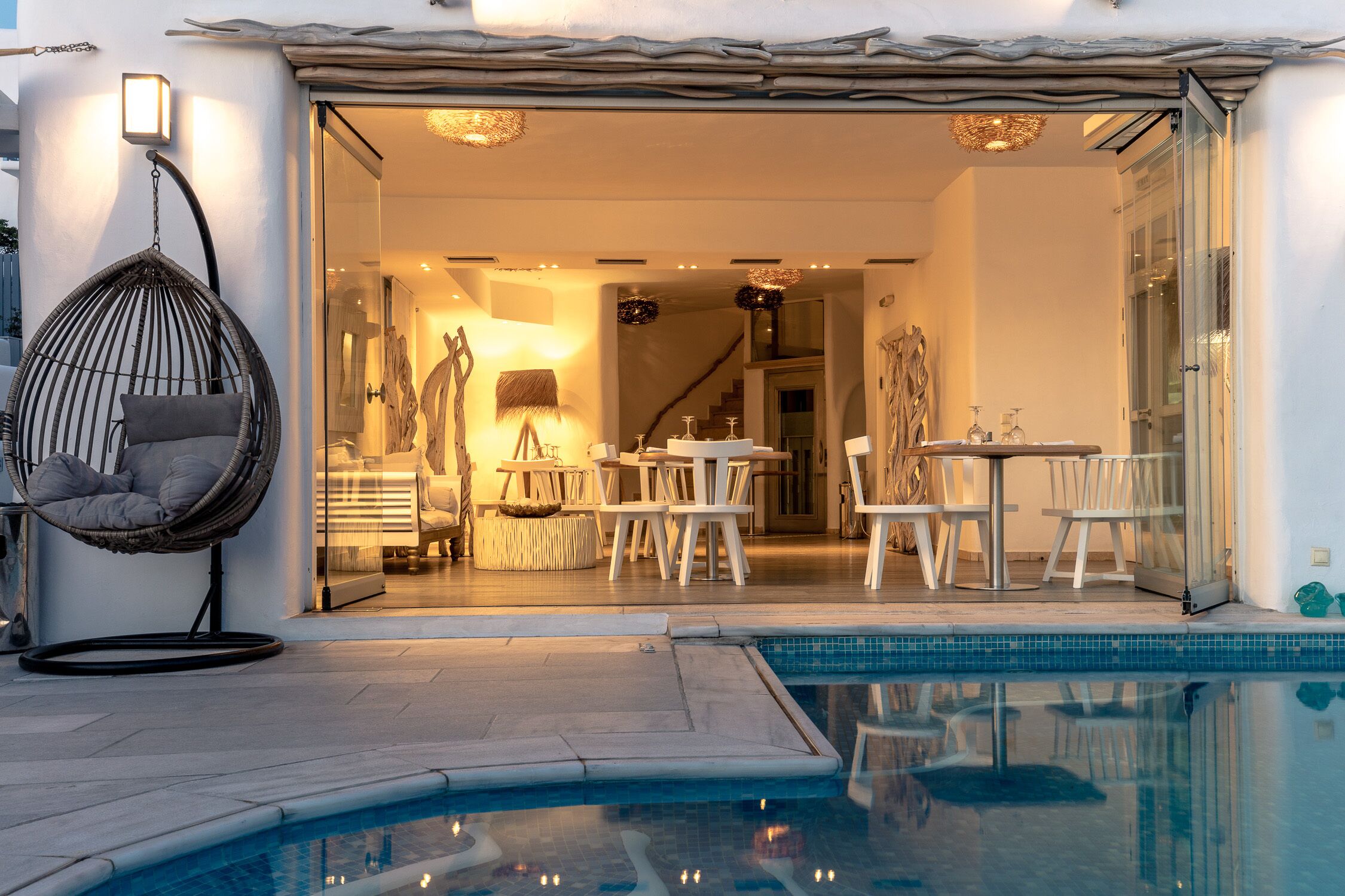 A modern outdoor pool area with a hanging chair, a dining area with white furniture, and a staircase leading to an upper level.