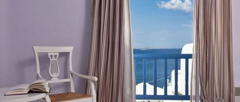 A room with a view of the sea, a white chair, a table with a book, a tray with a cup and glass, and striped curtains.