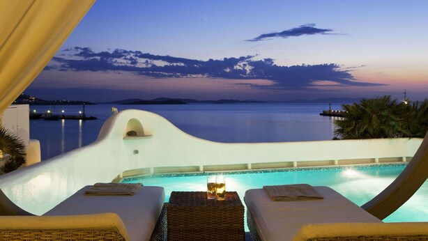Un área de descanso junto a la piscina con dos tumbonas, una mesa con bebidas y vista al mar al atardecer.