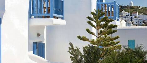 A white building with blue balconies and doors, wooden chairs, and a palm tree.
