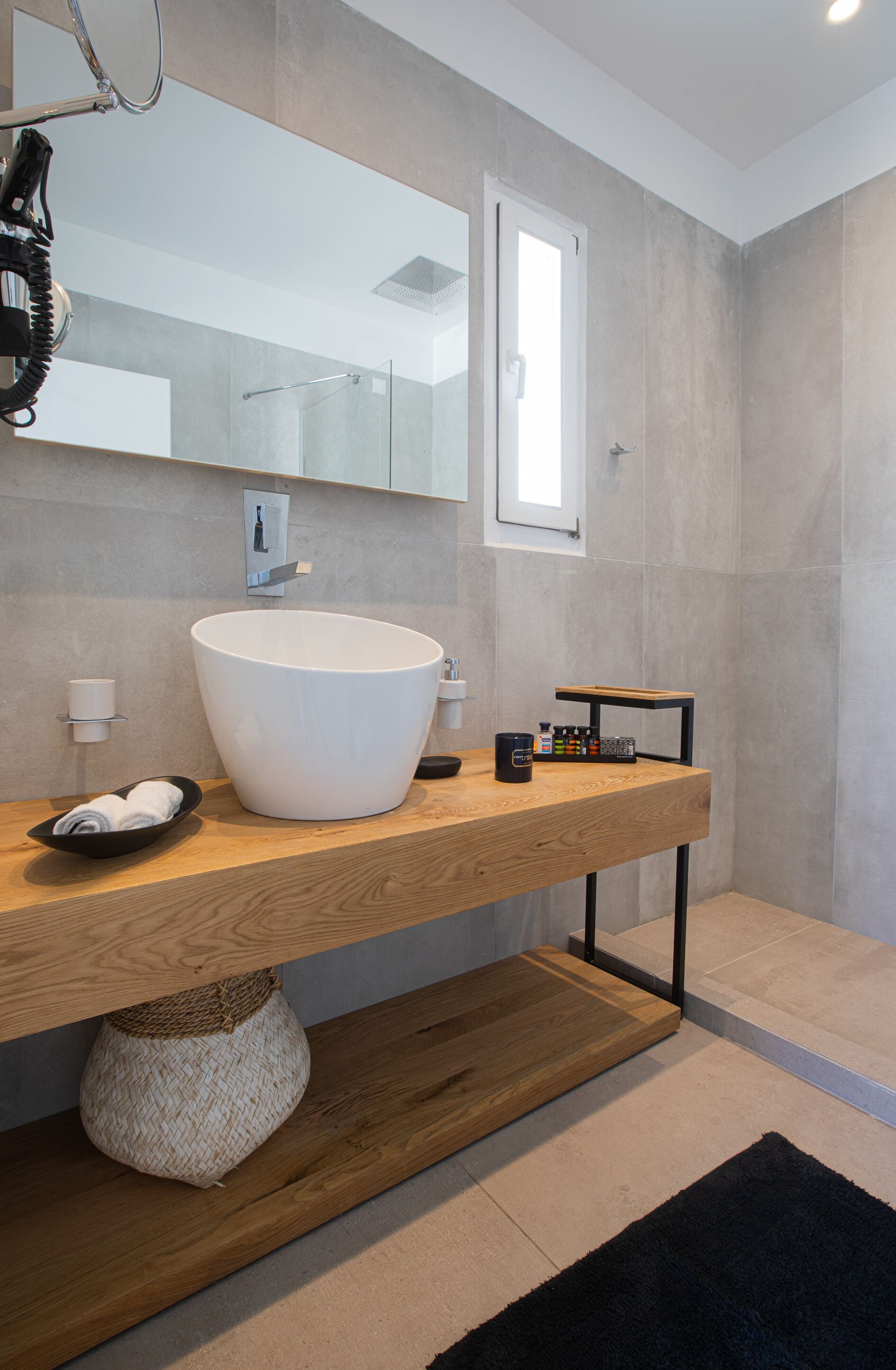 Un baño moderno con un lavabo blanco grande, tocador de madera y una pequeña estantería con artículos de aseo.