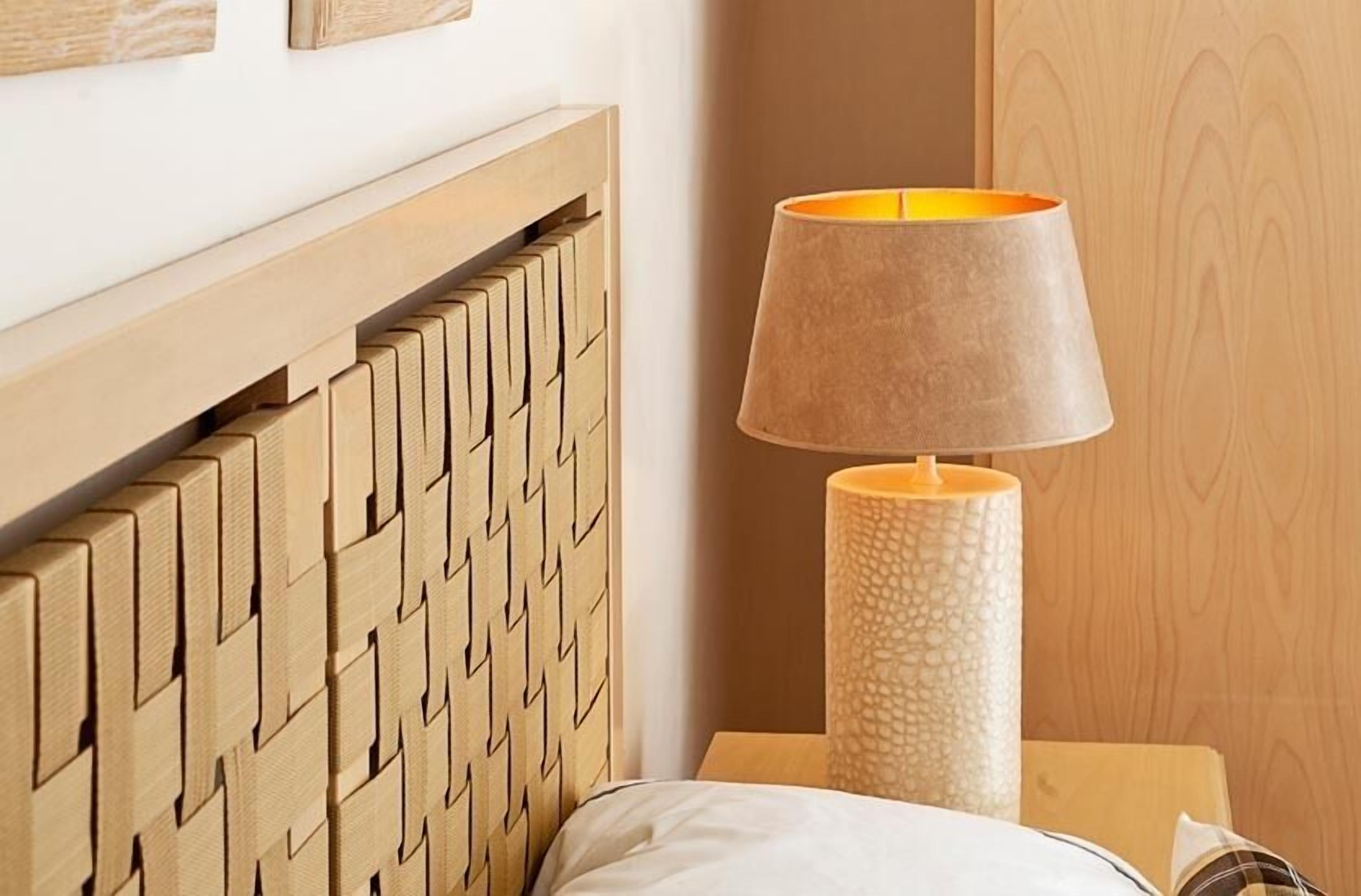 A wooden headboard with a geometric pattern, a textured cylindrical lamp, and a wooden wall with a subtle wood grain design.