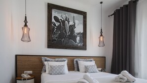 Una habitación de hotel con cama, dos lámparas de noche, teléfono, cuadro con motivos de cactus y ventana con cortinas.