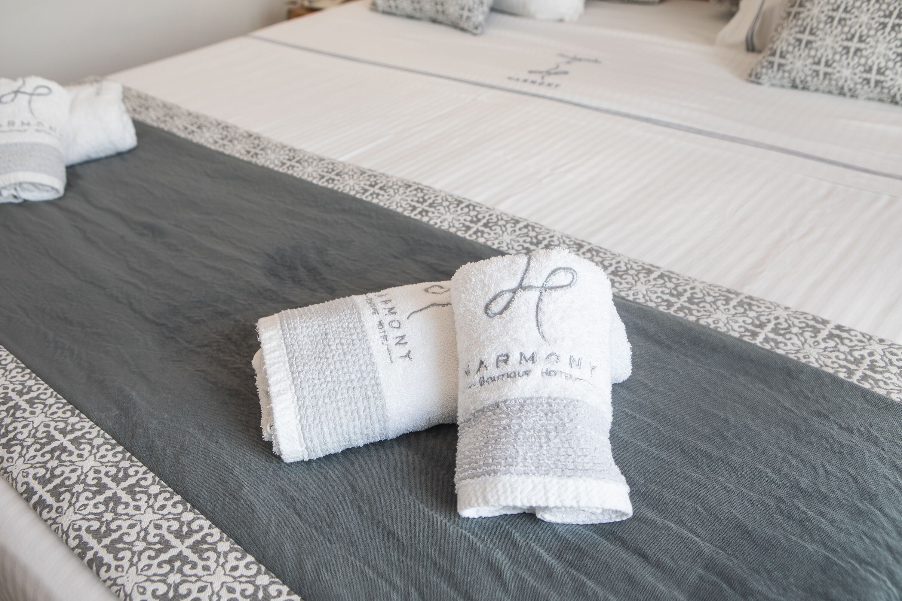 A neatly made bed with a grey and white patterned bedspread, two rolled towels with 'HARMONY' branding, and a patterned pillow.
