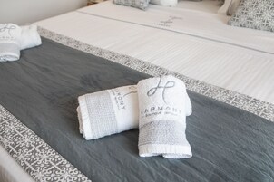 Una cama bien tendida con una colcha gris y blanca estampada, dos toallas enrolladas con la marca 'HARMONY' y una almohada estampada.