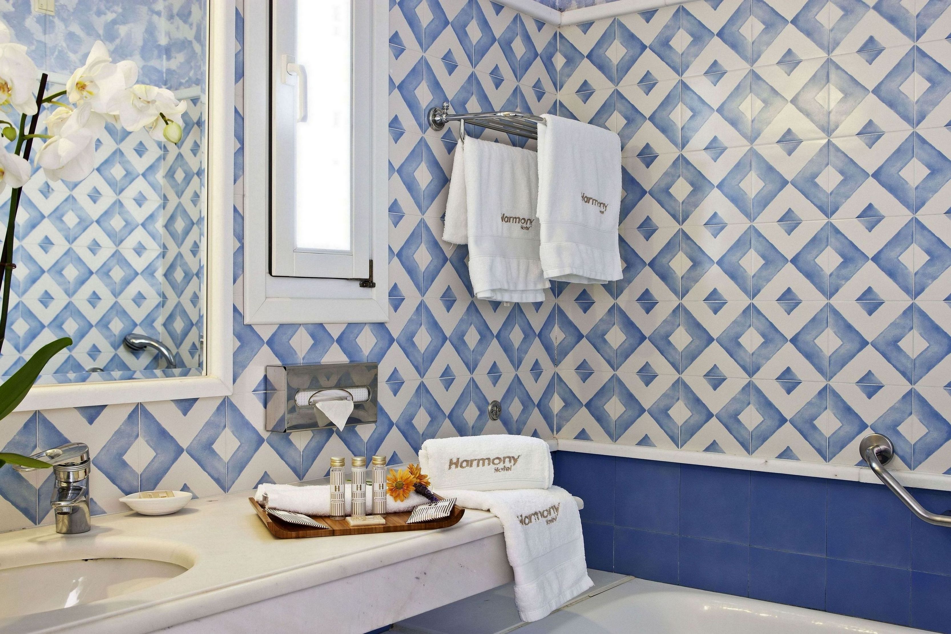 A bathroom with a blue and white diamond-patterned tile backsplash, a white sink, a mirror, and a bathtub. There are towels hanging on a rack and a tray with toiletries on the sink.