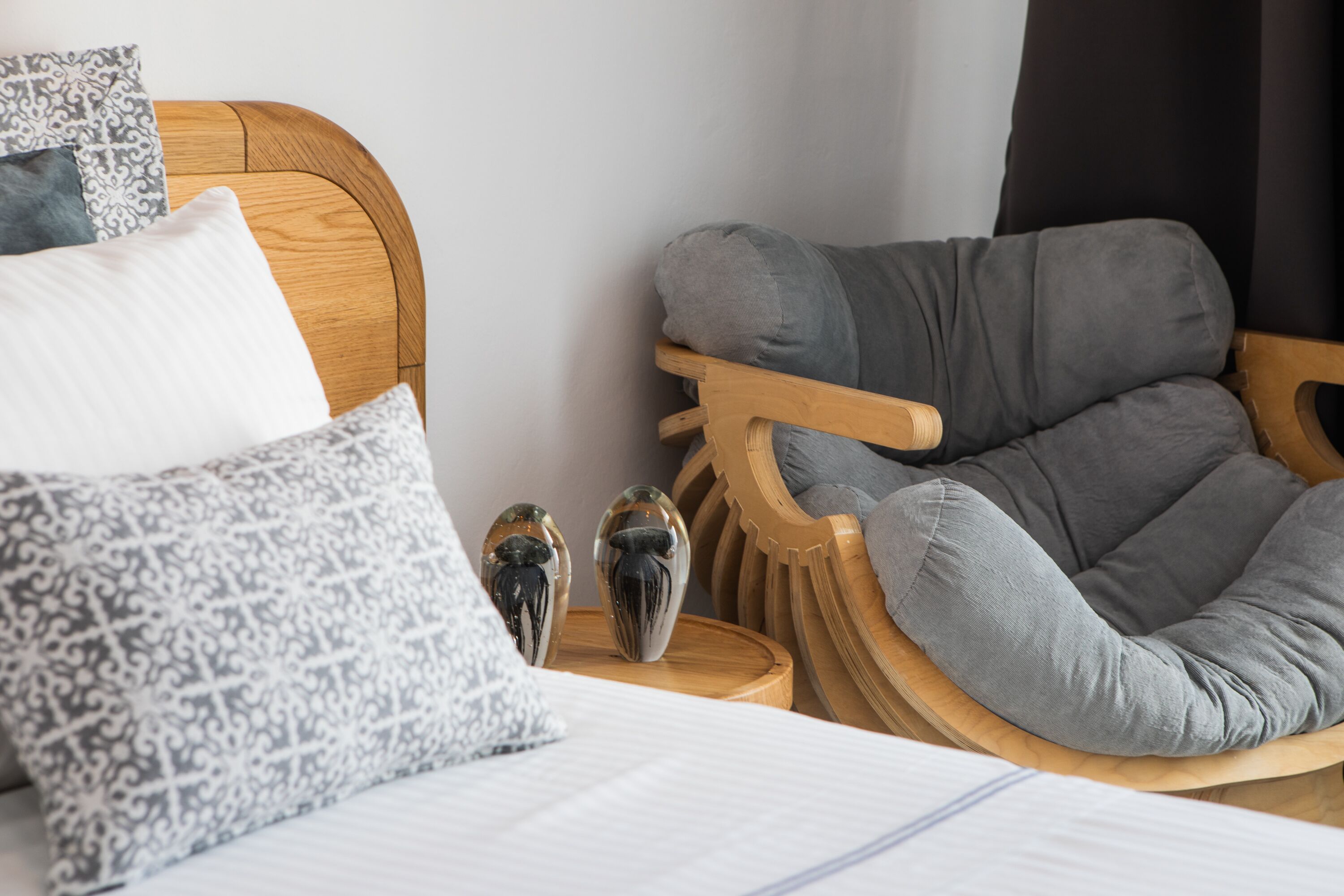 Une chambre à coucher bien aménagée, avec une tête de lit en bois, un fauteuil recouvert d’un tissu gris et des coussins à motifs.