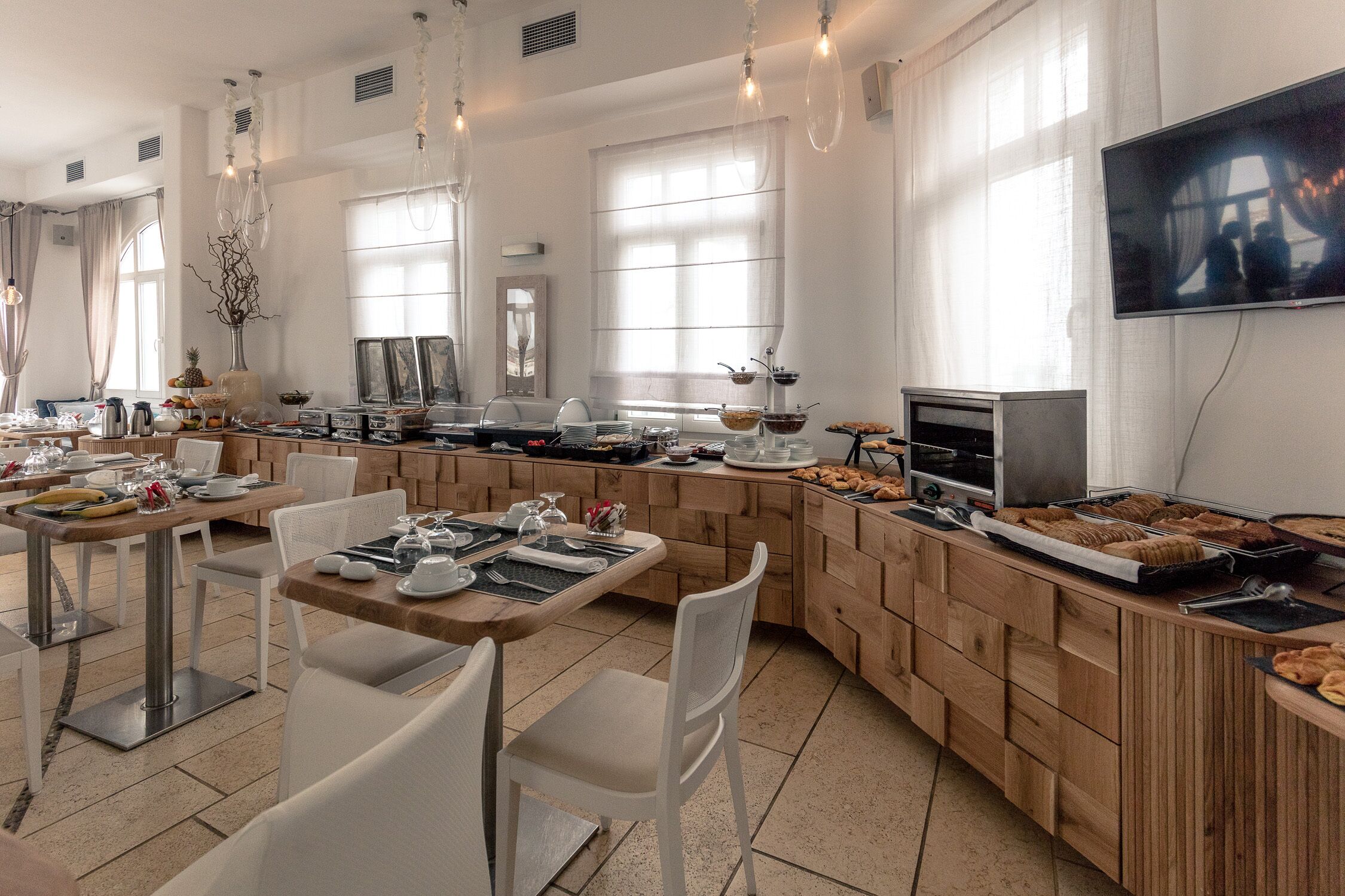 A modern dining area with wooden tables and chairs, a buffet counter with various food items, and a television mounted on the wall.
