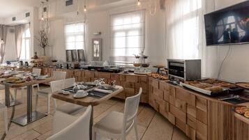 A modern dining area with wooden tables and chairs, a buffet counter with various food items, and a television mounted on the wall.