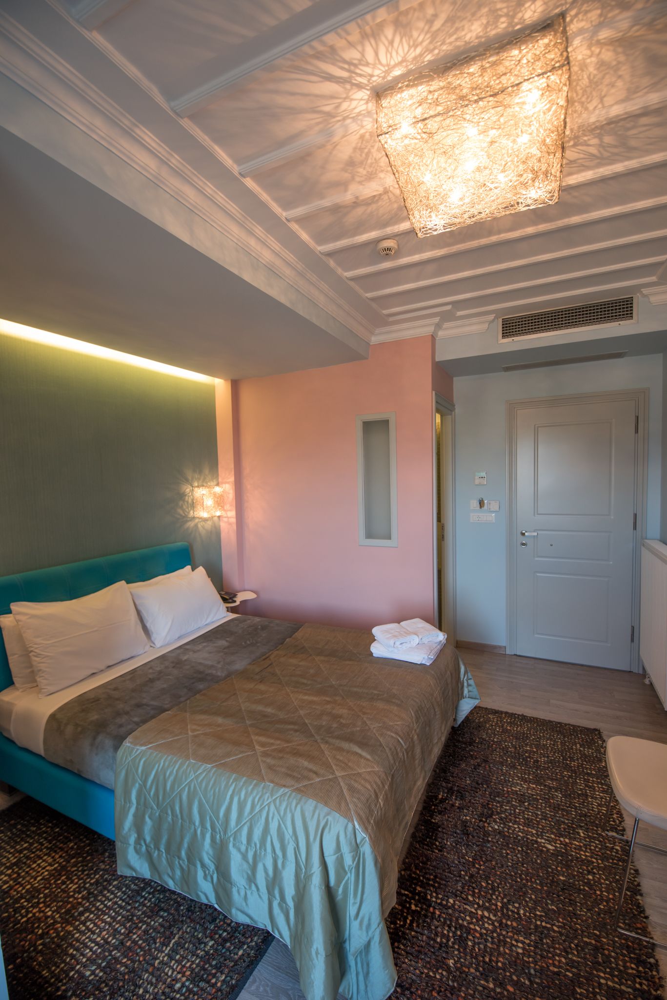 Une chambre d’hôtel comprenant un lit, une table de chevet, une lampe fixée au mur, une porte et un luminaire au plafond.