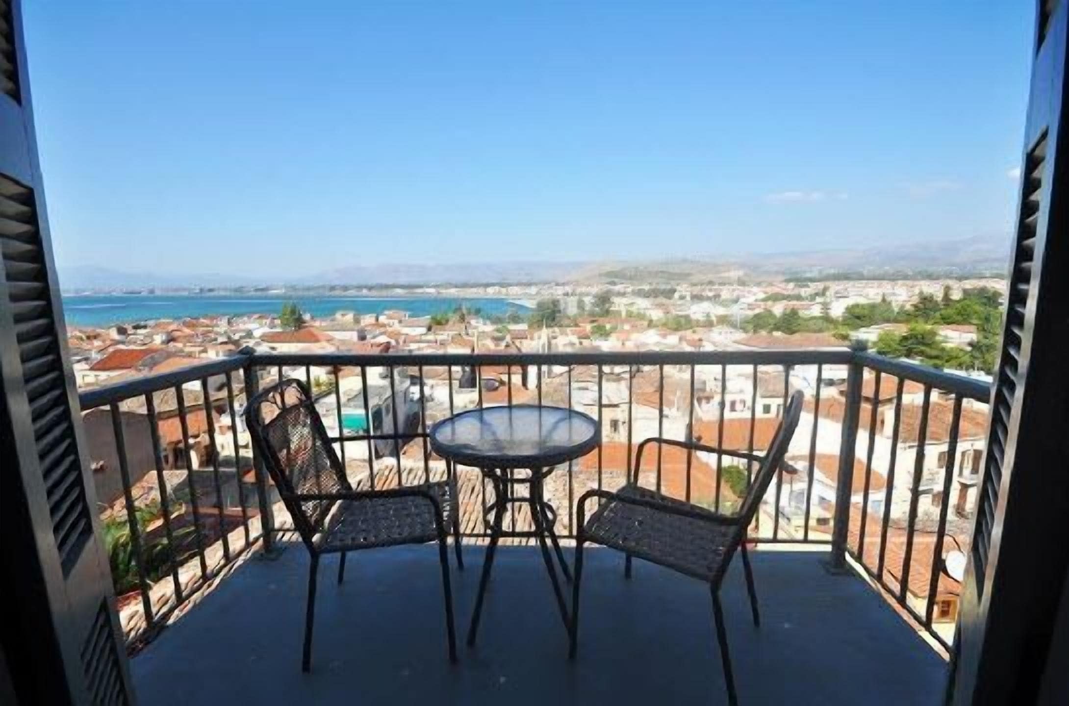 Un balcón con una mesa y dos sillas, con vista a una ciudad costera con edificios y una masa de agua.