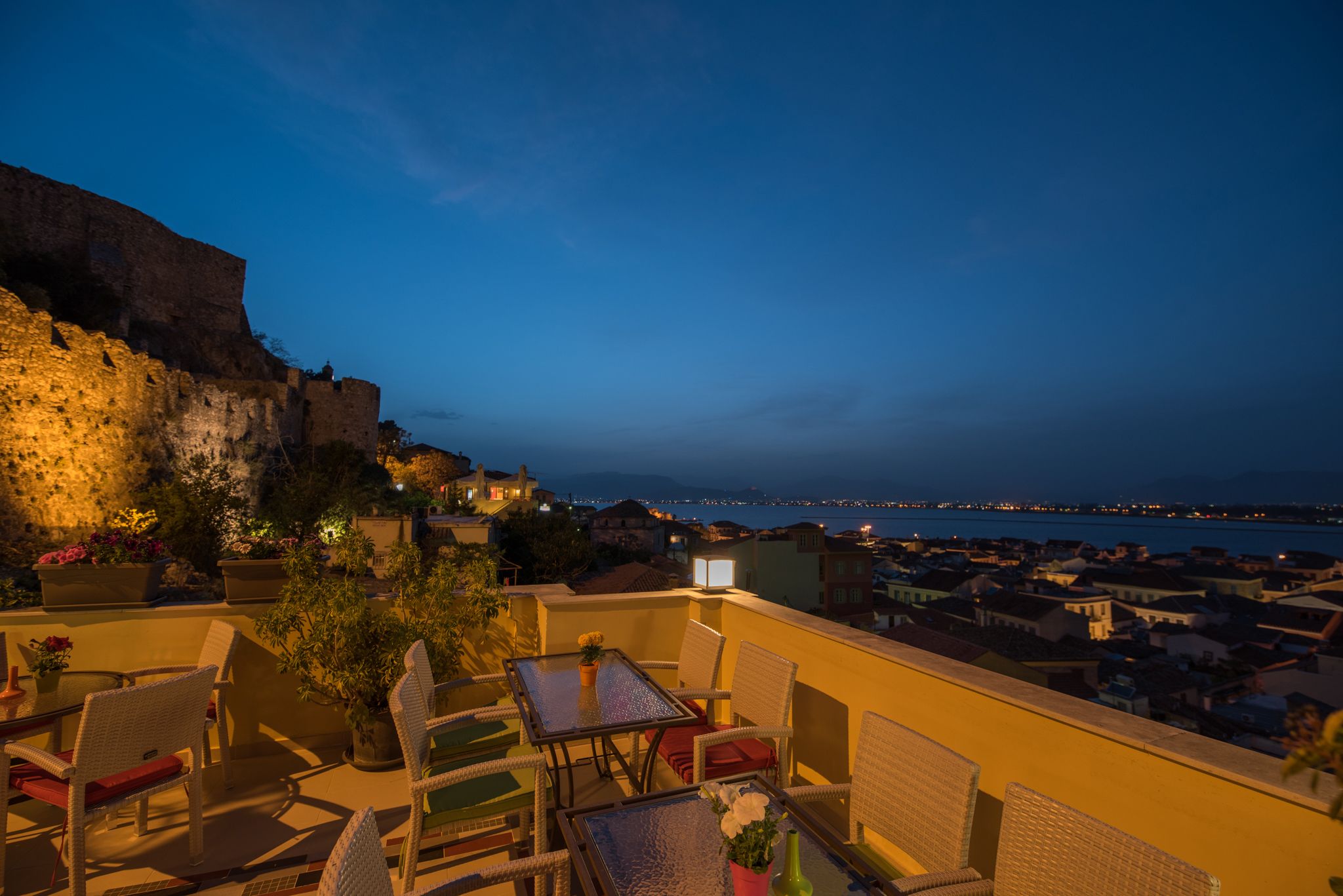 Une terrasse meublée de mobilier en osier, avec une table, d’où l’on peut admirer le paysage urbain de la côte au crépuscule.