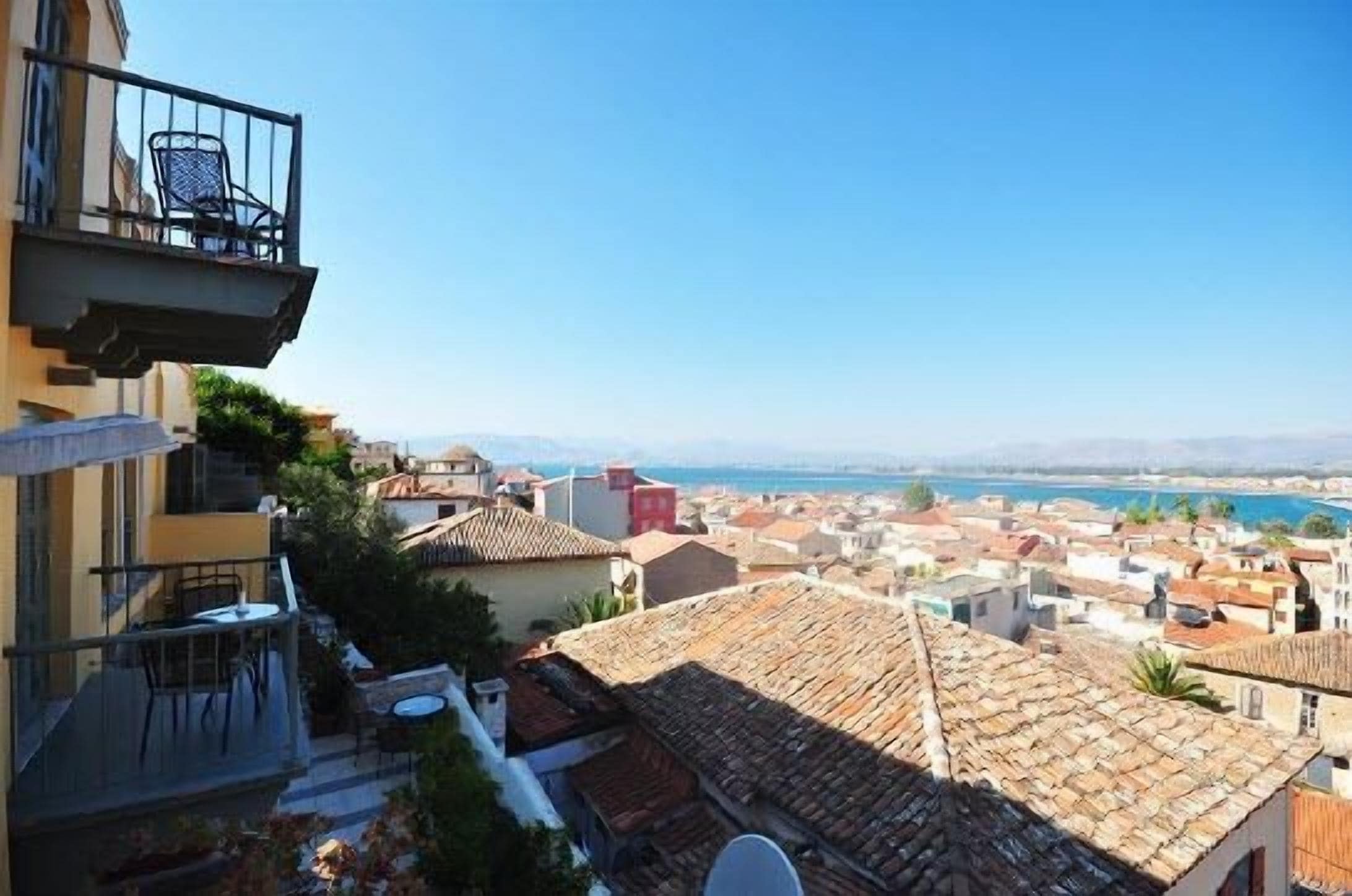 A balcony with a chair and table, overlooking a coastal town with terracotta roofs and a clear blue sky.