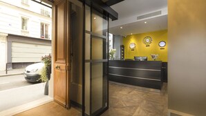 A hotel reception area with a glass door entrance, a reception desk, and two wall clocks. - Hôtel International Paris (Paris)