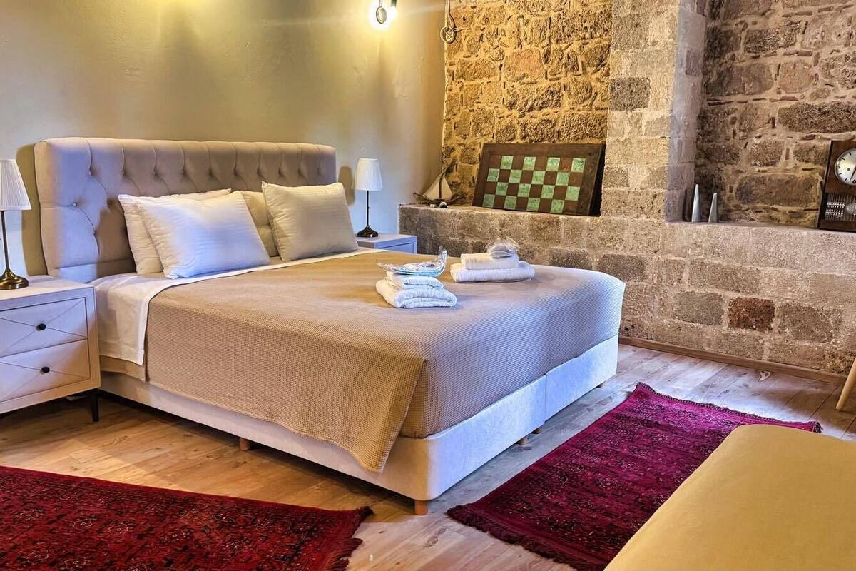 A bedroom with a stone wall, a bed with a headboard, a nightstand, a lamp, a clock, and a checkered wall decoration.