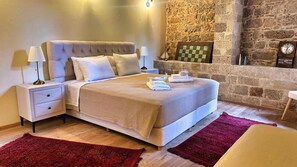 A bedroom with a stone wall, a bed with a headboard, a nightstand, a lamp, a clock, and a checkered wall decoration. - Avalon Boutique Hotel Rhodes (Rhodes)