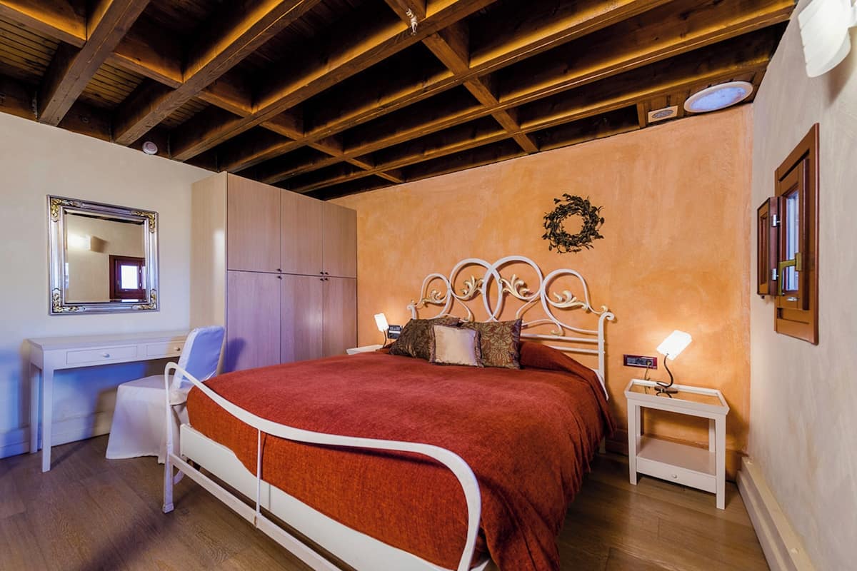 A bedroom with a wooden ceiling, a bed with a white metal frame, a bedside table with a lamp, a wardrobe, and a mirror on the wall.