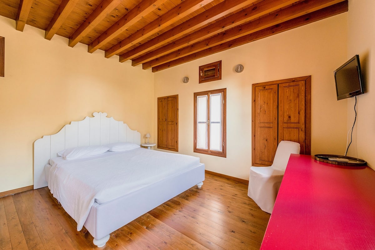 A bedroom with a wooden ceiling, a bed, a chair, and a TV mounted on the wall.