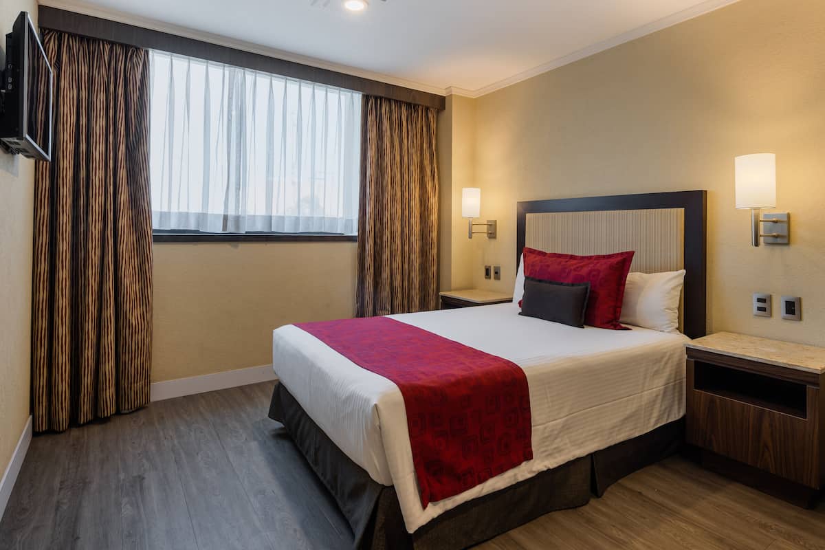 A hotel room with a large bed, a TV mounted on the wall, a bedside table, and a window with curtains.