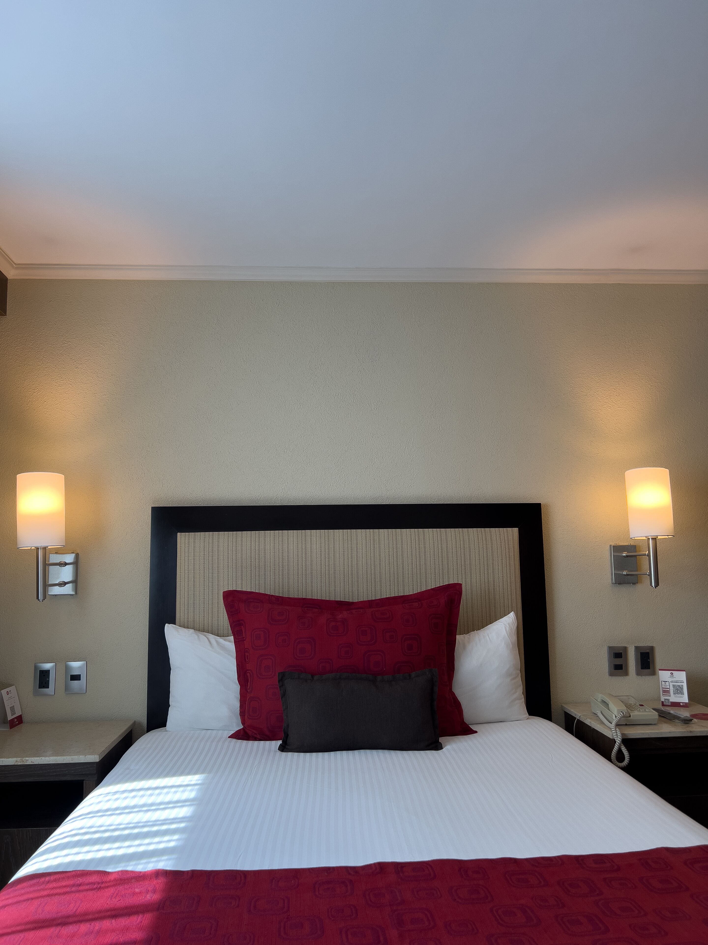 A hotel room with a large bed, two bedside lamps, a telephone, and a wall-mounted light switch.