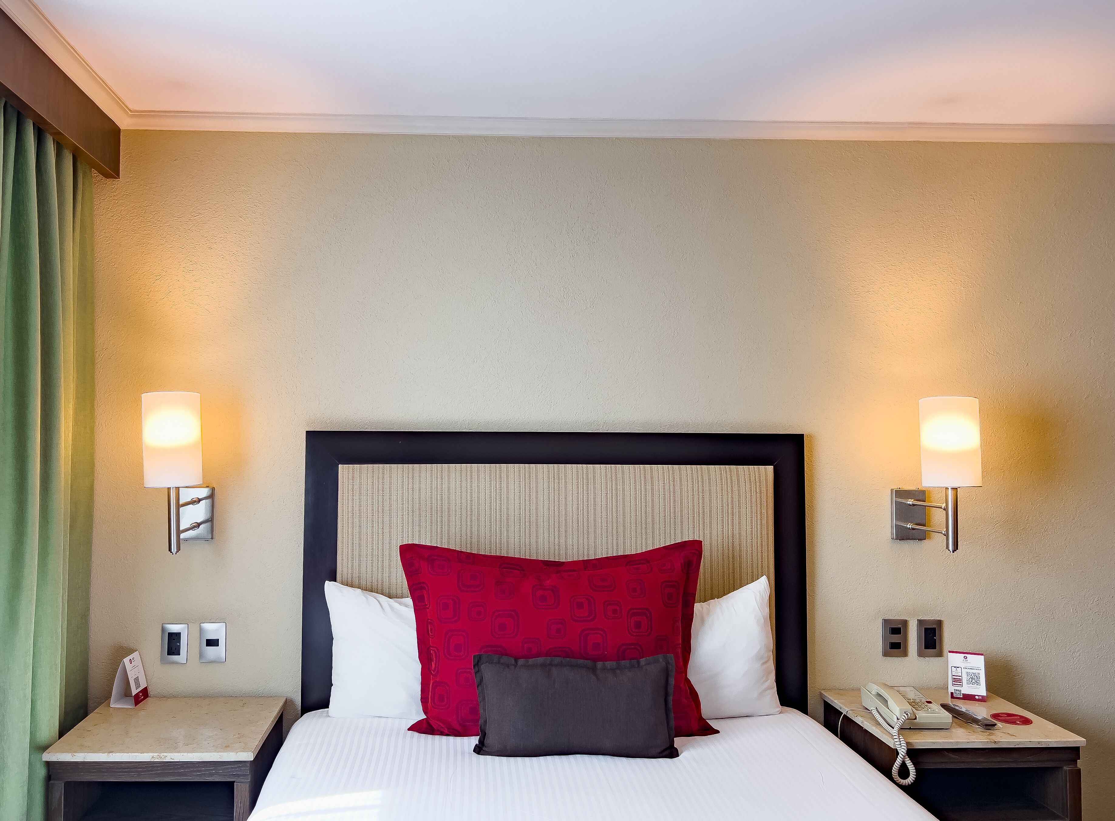 Habitación de hotel con cama, dos mesitas de noche, teléfono y lámpara de pared.