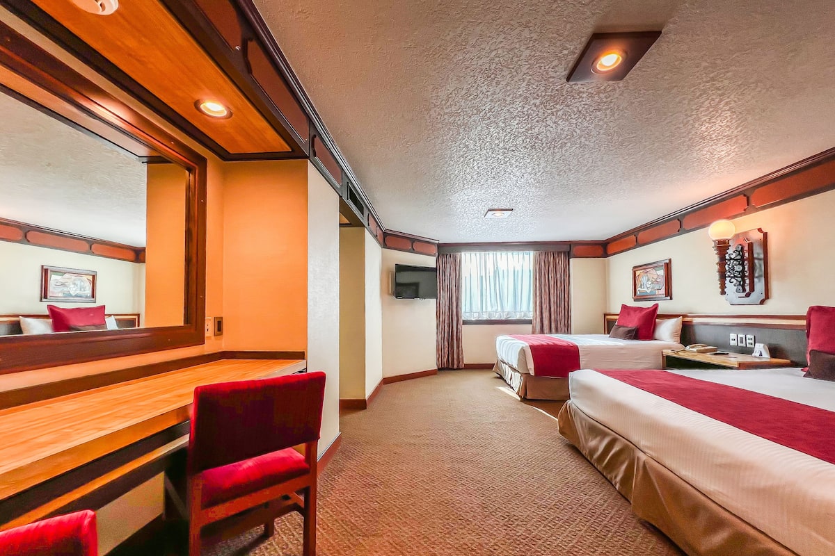 A hotel room with a wooden desk, two beds, a television, and a window with curtains.